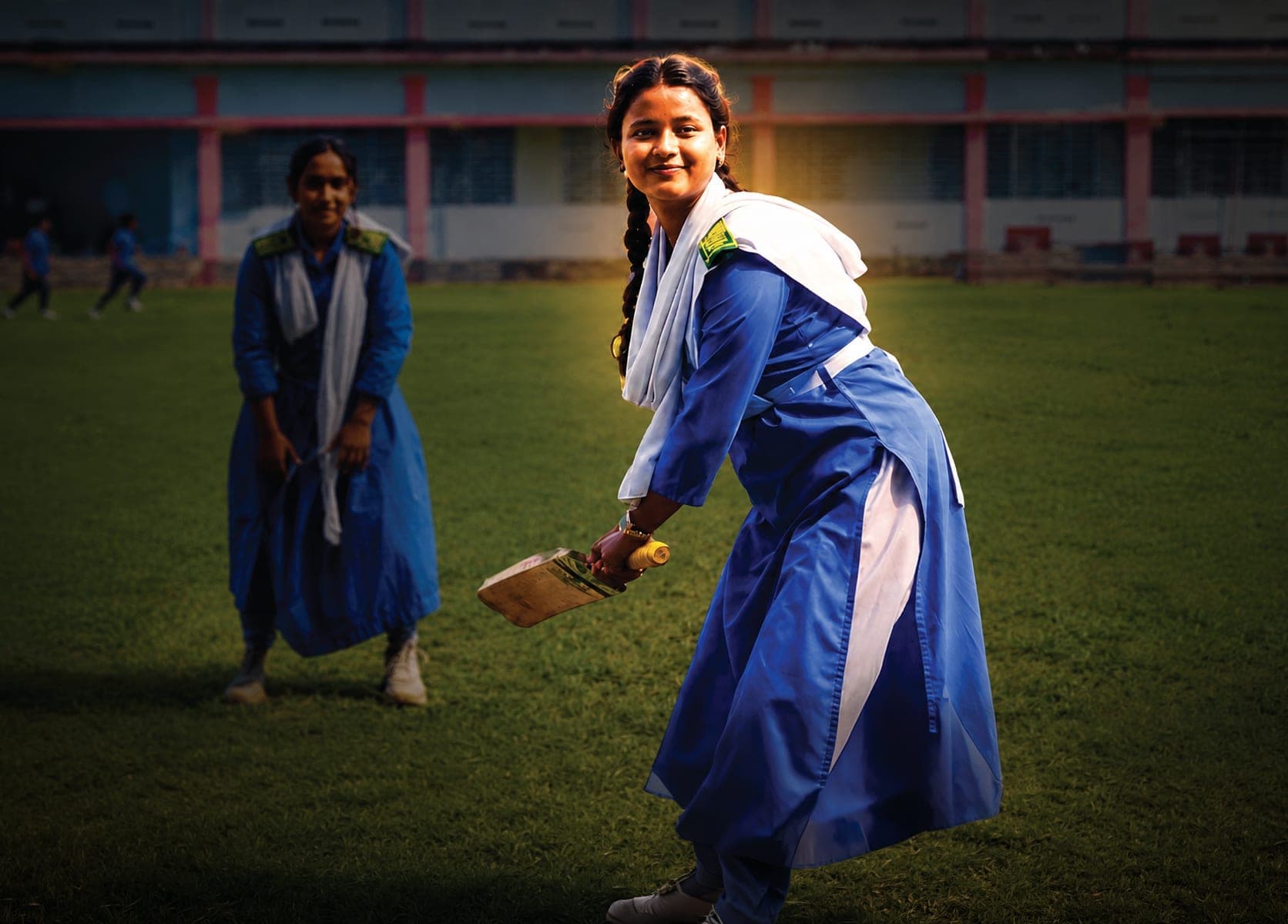 A schoolgirl in a blue and white uniform holds a cricket bat mid-play on a school field, smiling towards the camera, with a classmate behind her.