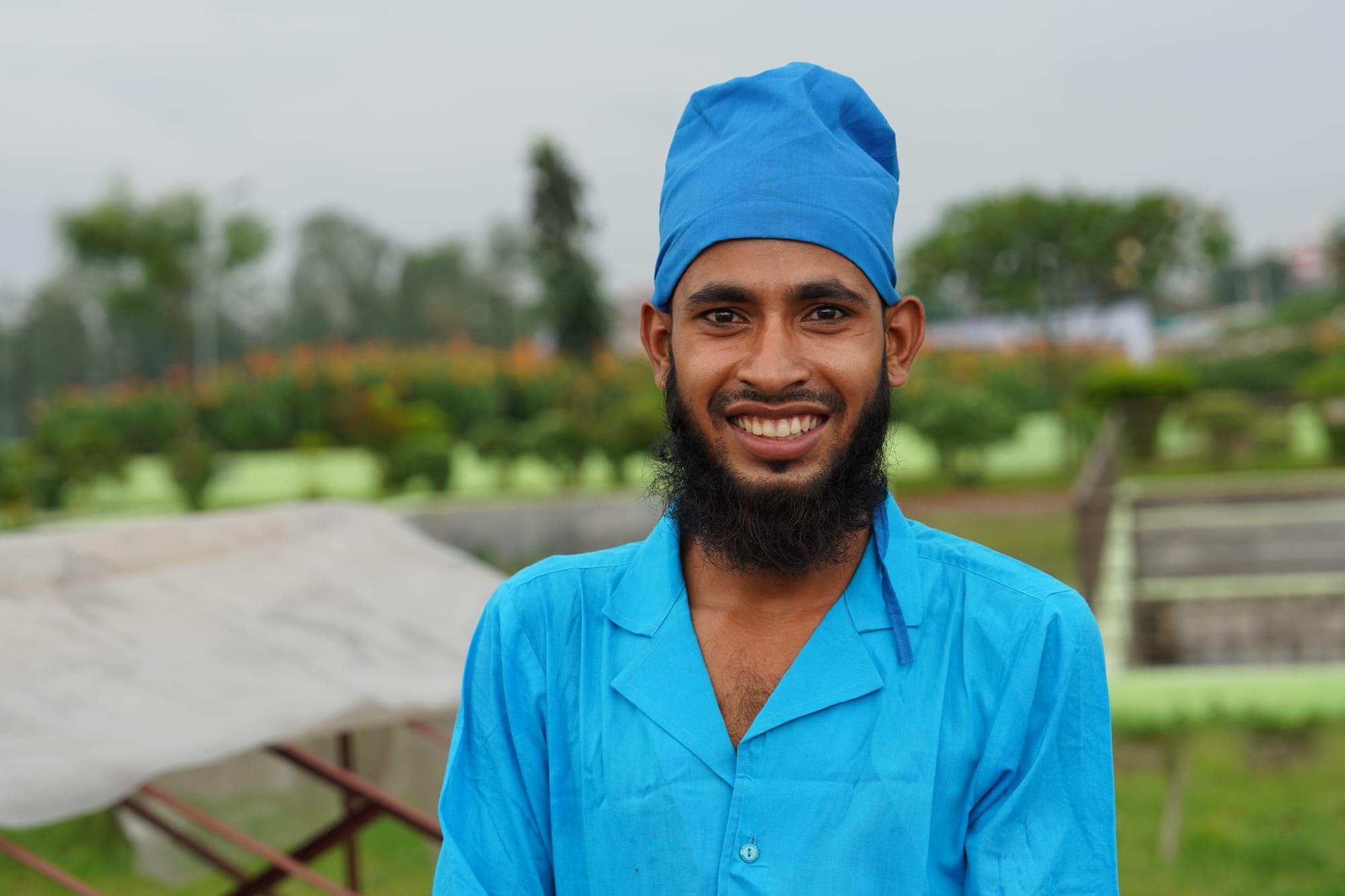 Azad Rahman smiles at the camera in a bright blue work uniform and cap, standing in the grounds of the faecal sludge treatment plant.