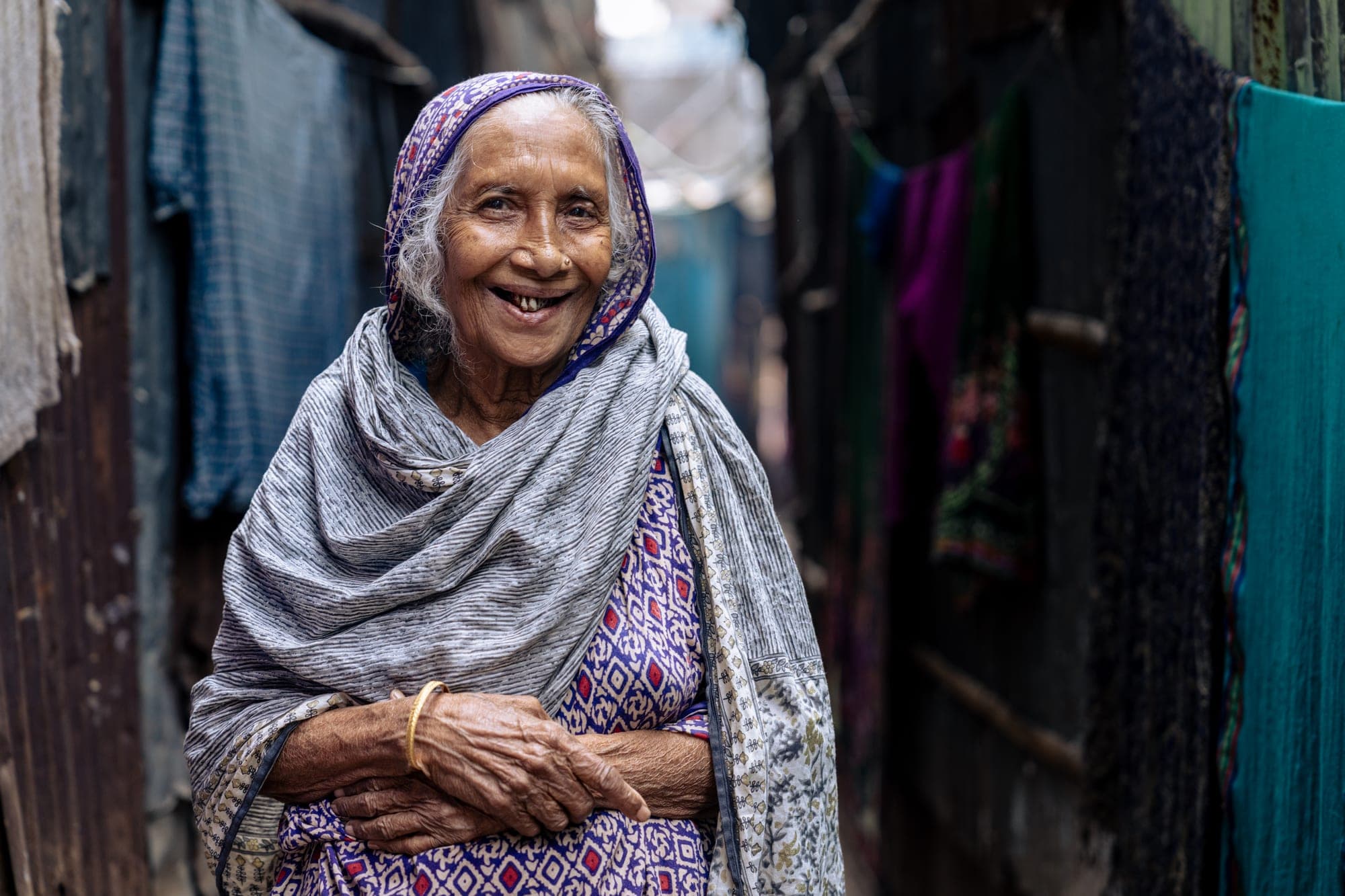 Surja Banu, an older woman in a patterned sari and grey shawl, laughs warmly in the narrow lane of her settlement.