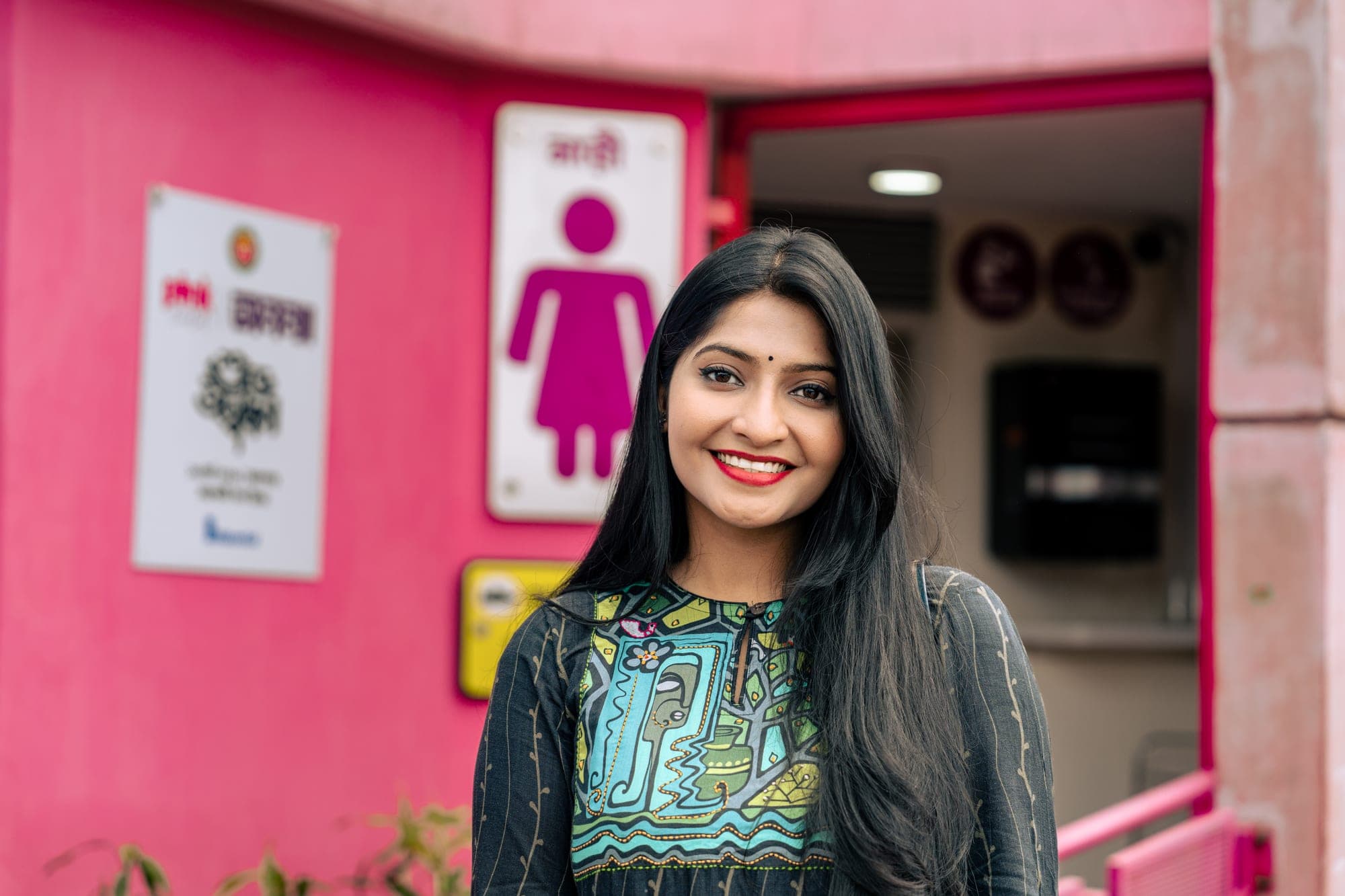 A young woman smiles outside Ananya, a bright pink public toilet for women, with its women’s pictogram sign visible behind her.