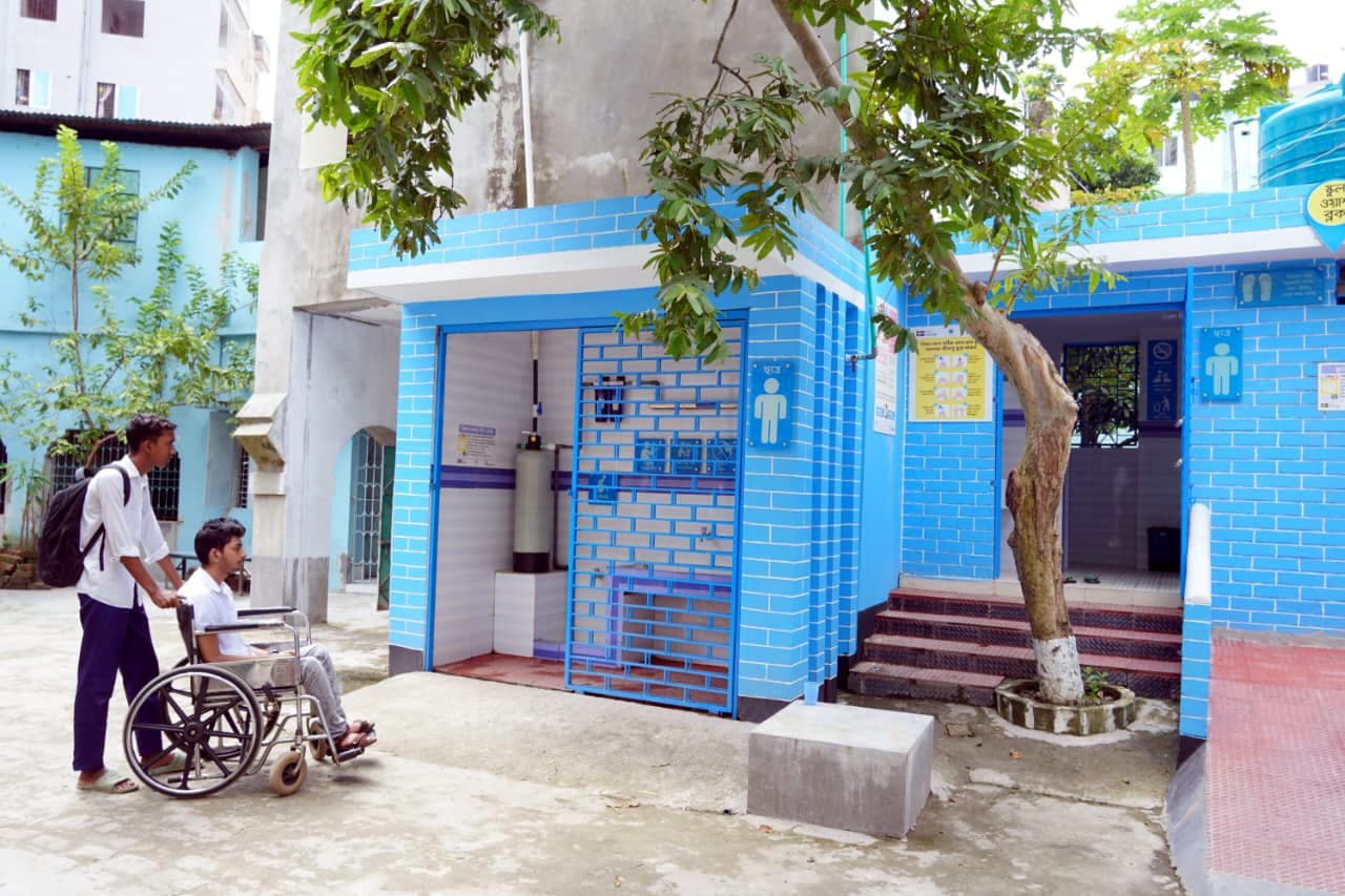 A student using a wheelchair is accompanied by a classmate towards a newly built, blue-tiled accessible school washroom and drinking water block.